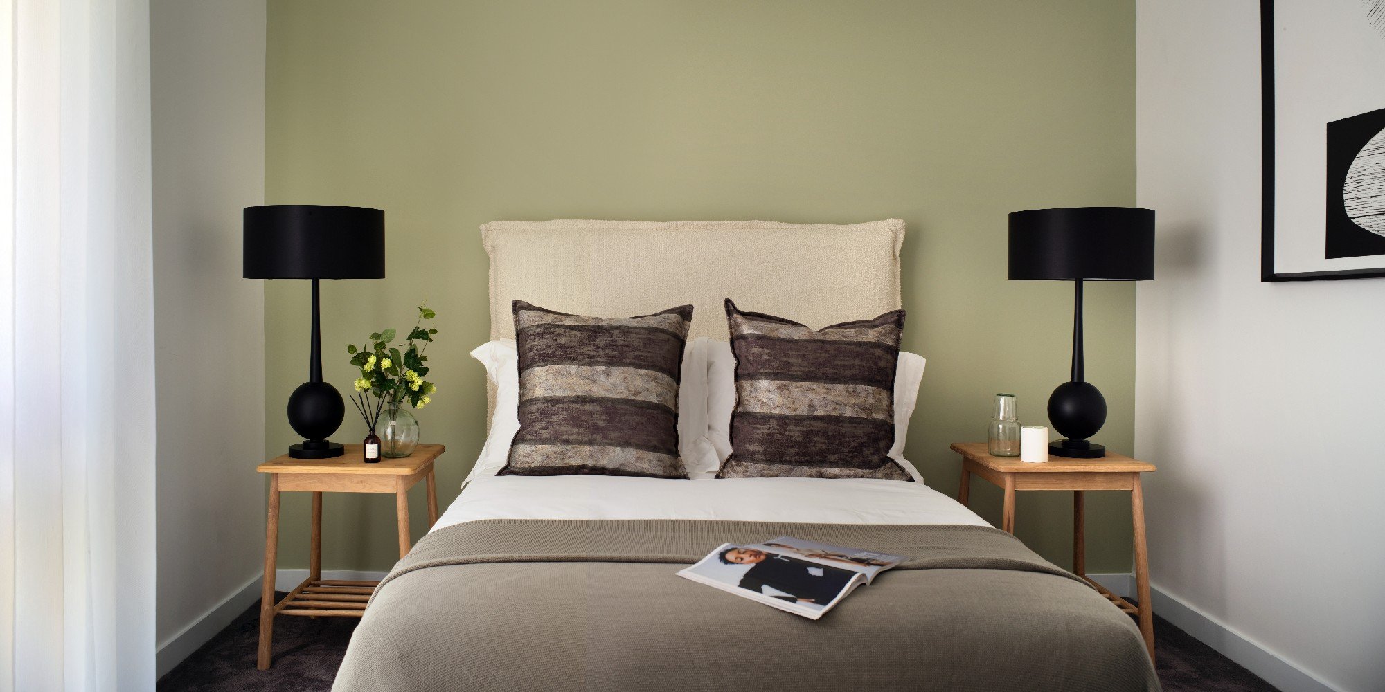 A double bed against a green wall with neutral toned cushions and duvet, with an open magazine on the bed