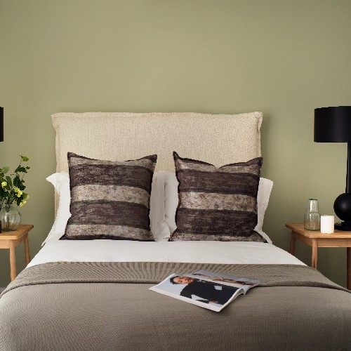 A double bed against a green wall with neutral toned cushions and duvet, with an open magazine on the bed