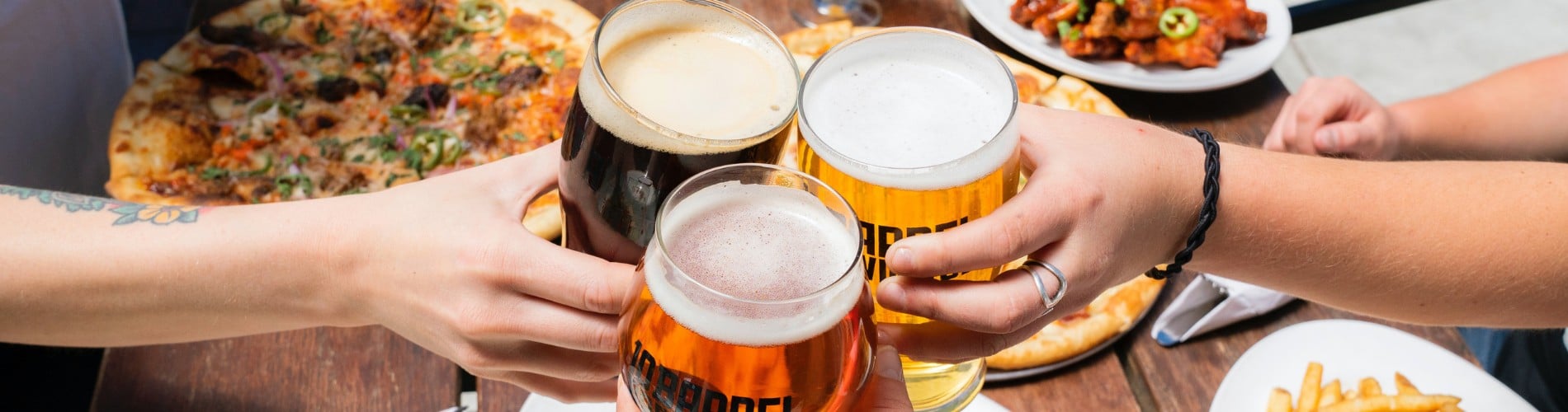 Three people holding pints of beer over a table of pizzas