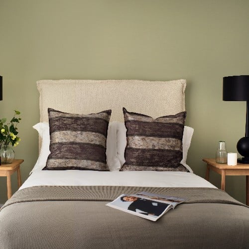 A double bed against a green wall with neutral toned cushions and duvet, with an open magazine on the bed